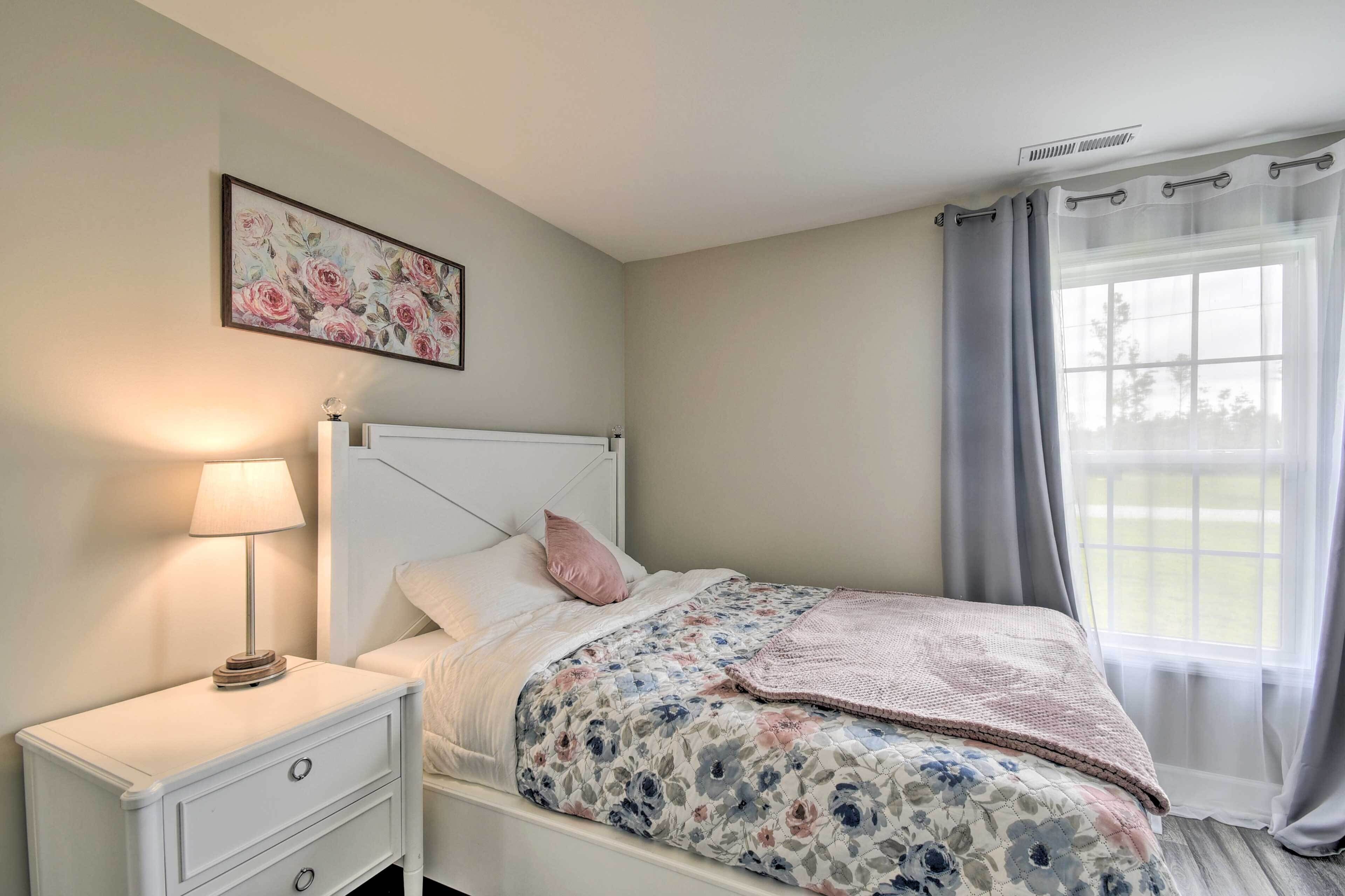 A bedroom with a bed, a nightstand with a lamp, a framed floral artwork on the wall, and a window with curtains.