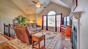 A spacious living room with a high ceiling, a fireplace, a large window with curtains, a ceiling fan, and a television.