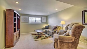 A living room with a brown armchair, a sofa, a coffee table, and a wooden cabinet. There is a window with blinds and a lamp on a side table.