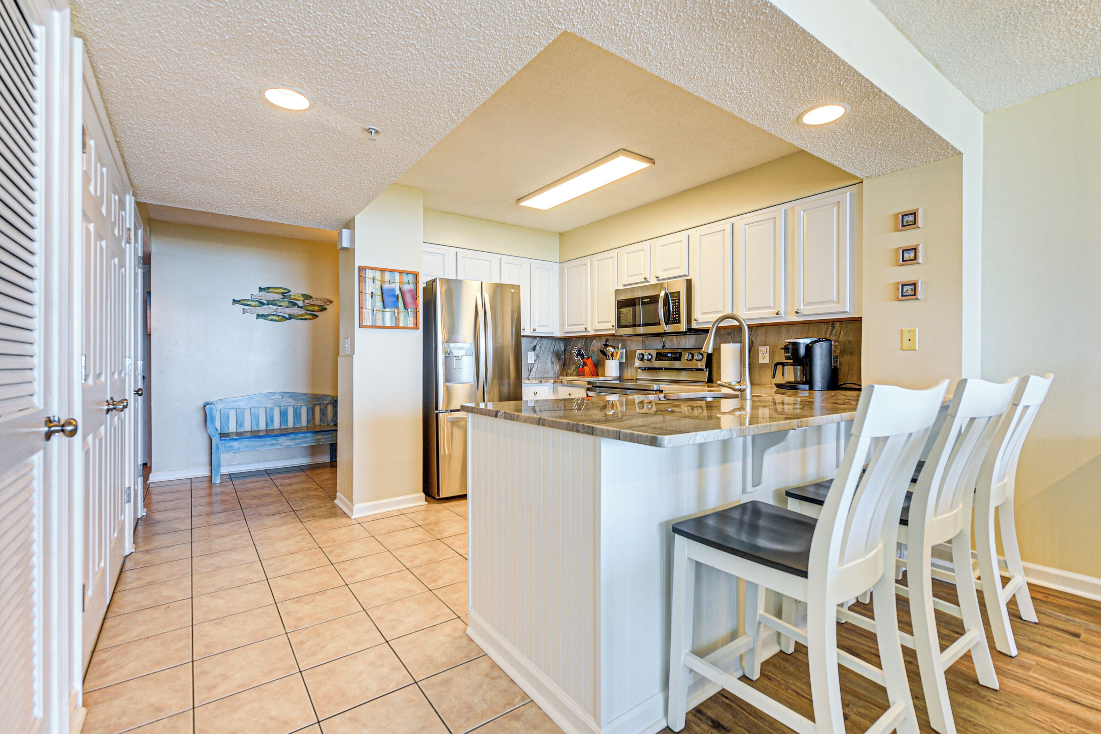 A kitchen with white cabinets, a stainless steel refrigerator, a microwave, and a coffee maker. There is a bench in the corner and a dining area with white chairs.