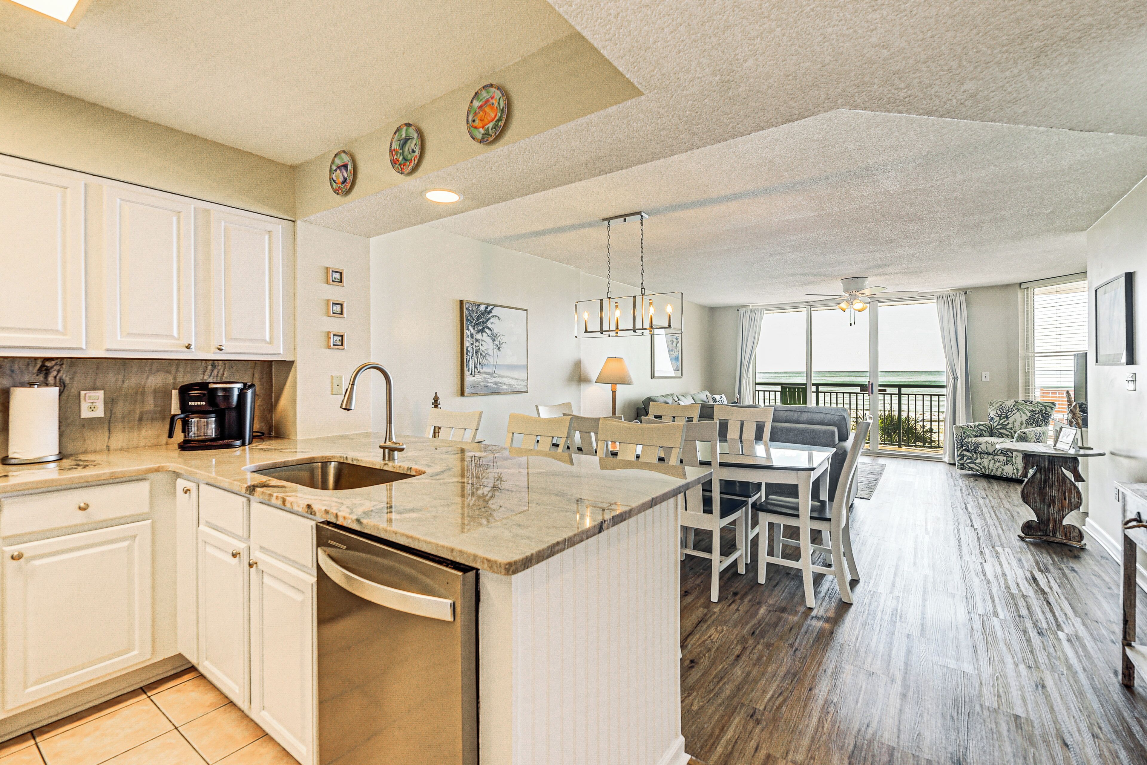 A spacious kitchen with white cabinets, a built-in dishwasher, and a large island. There is a dining area with a table and chairs, and a balcony with a view.