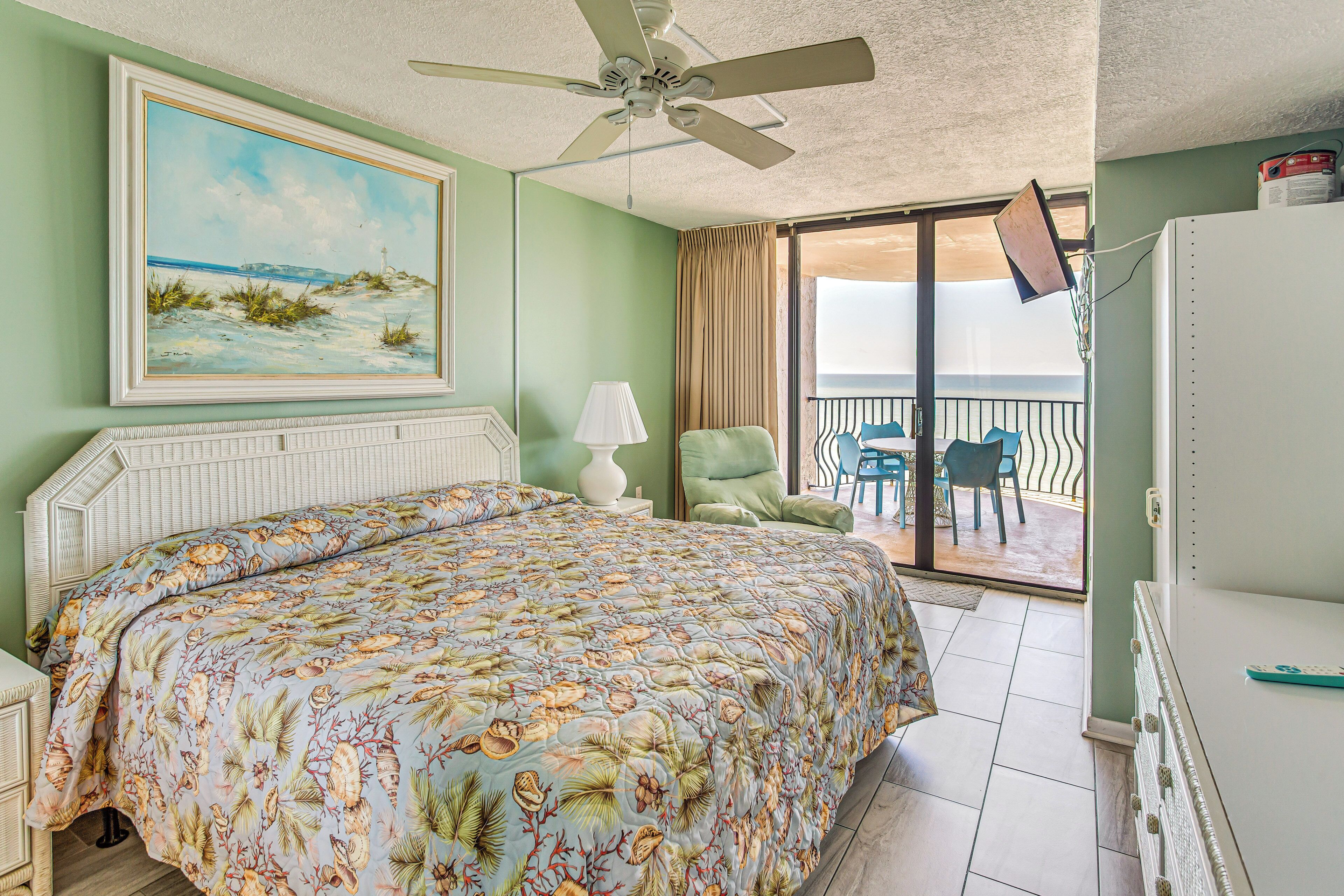 Ein Schlafzimmer mit einem Bett, einem Deckenventilator, einem Strandbild, einem Sessel und Meerblick durch eine Schiebetür.