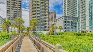 A wooden boardwalk leading to high-rise buildings with balconies, surrounded by lush greenery. - Oceanfront Condo w/ Balcony in Myrtle Beach! (Myrtle Beach)