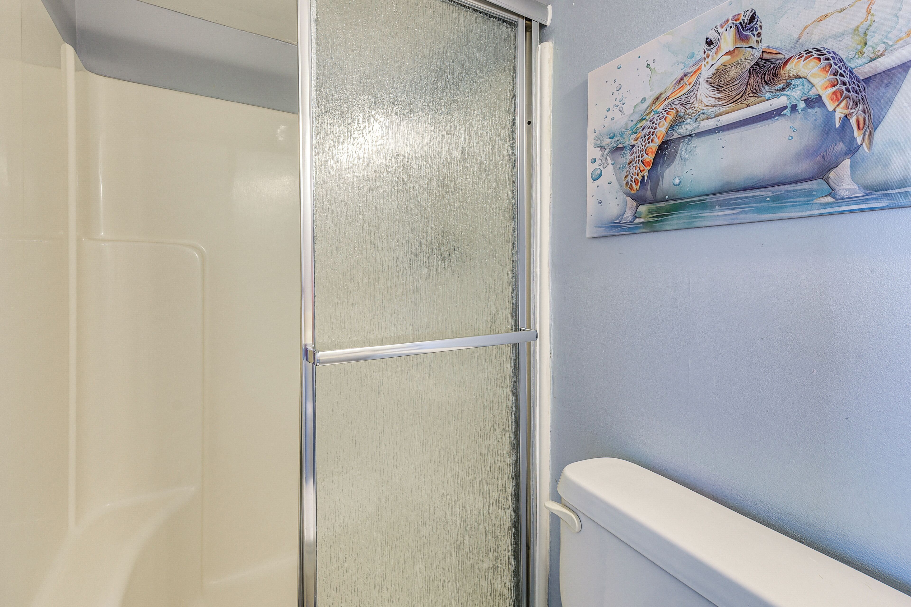 A bathroom with a glass shower enclosure, a white toilet, and a wall-mounted painting of a turtle in a bathtub.