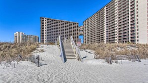 A beachfront hotel with a staircase leading to the entrance, surrounded by sand and sparse vegetation. - Resort Perks & Pools: Townhome By Barefoot Landing (Myrtle Beach)