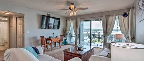 A living room with a ceiling fan, a TV mounted on the wall, a dining area with a table and chairs, and a view of the outdoors through large windows.