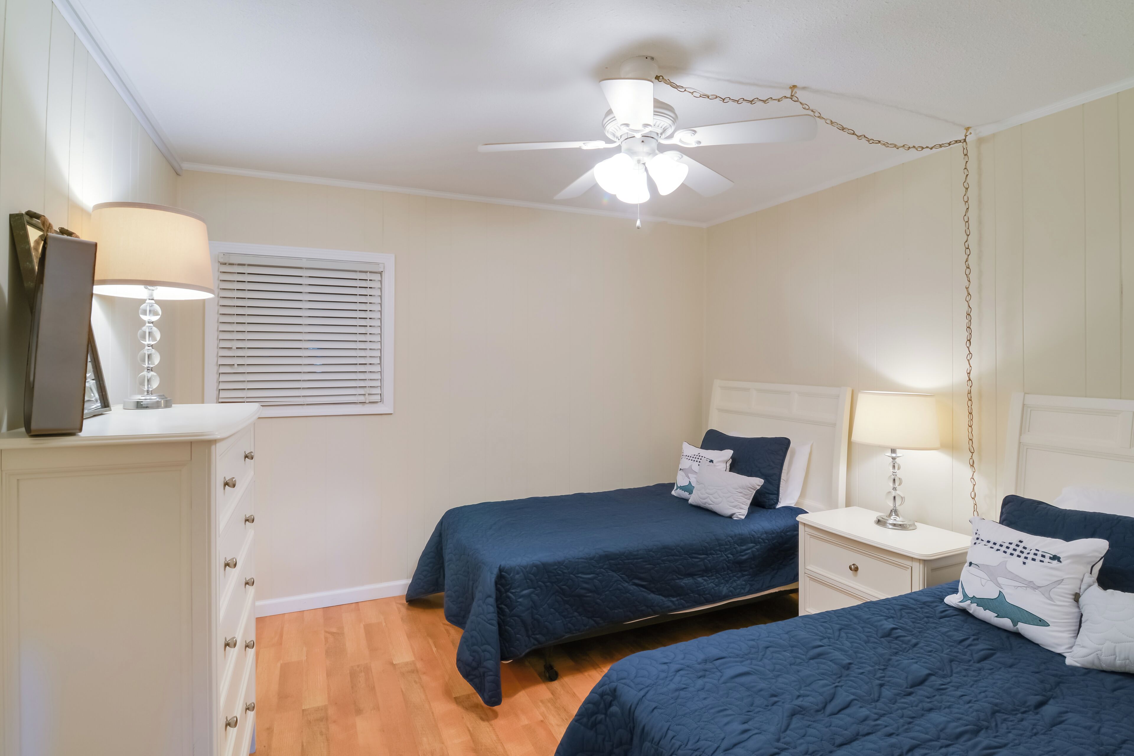 Ein Schlafzimmer mit zwei Betten, einem Deckenventilator, einem Nachttisch mit Lampe und einer Kommode.
