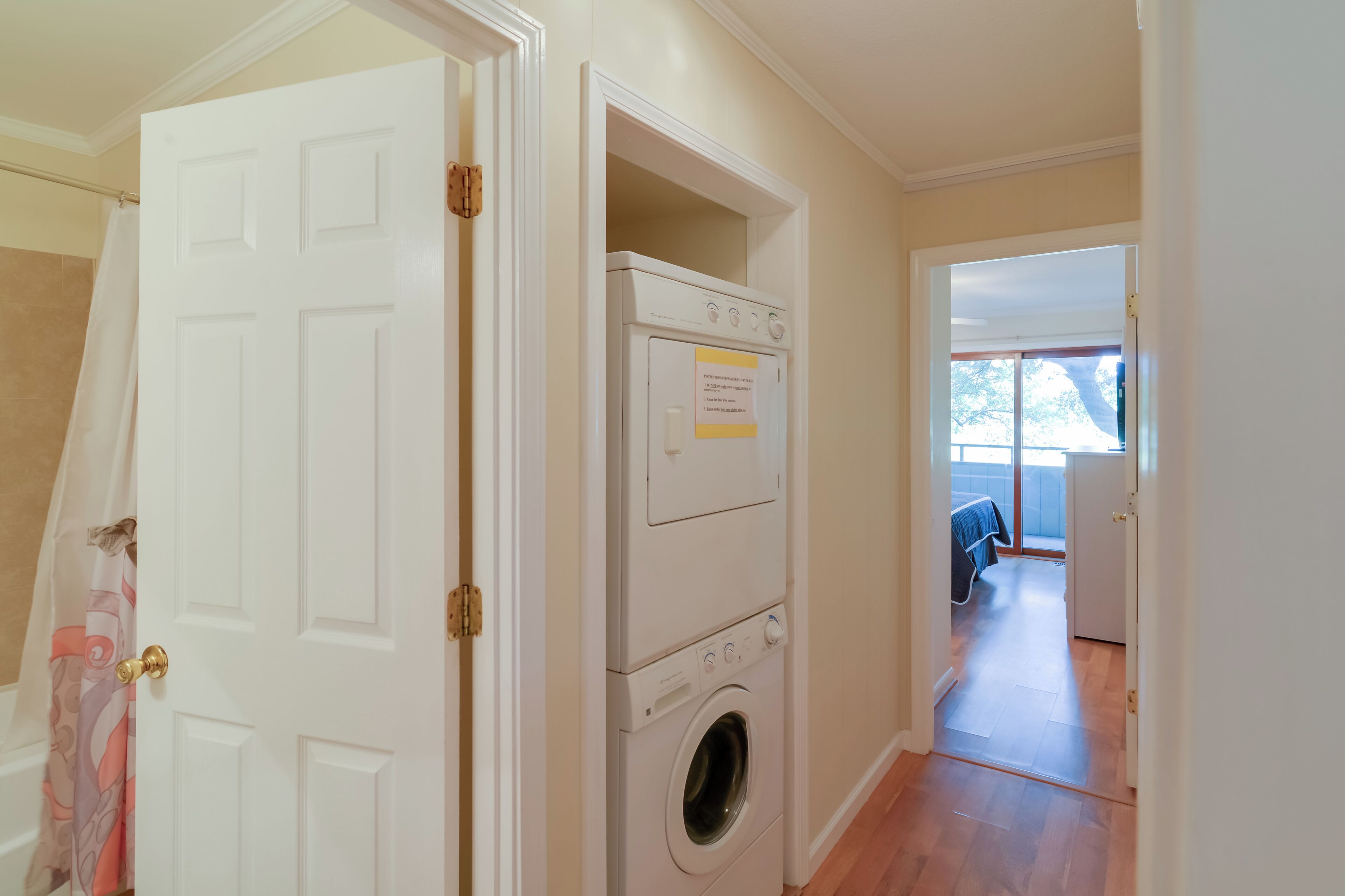 A laundry room with a front-loading washer and dryer, a door with a towel hanging, and a view into a bedroom with a bed and a window.