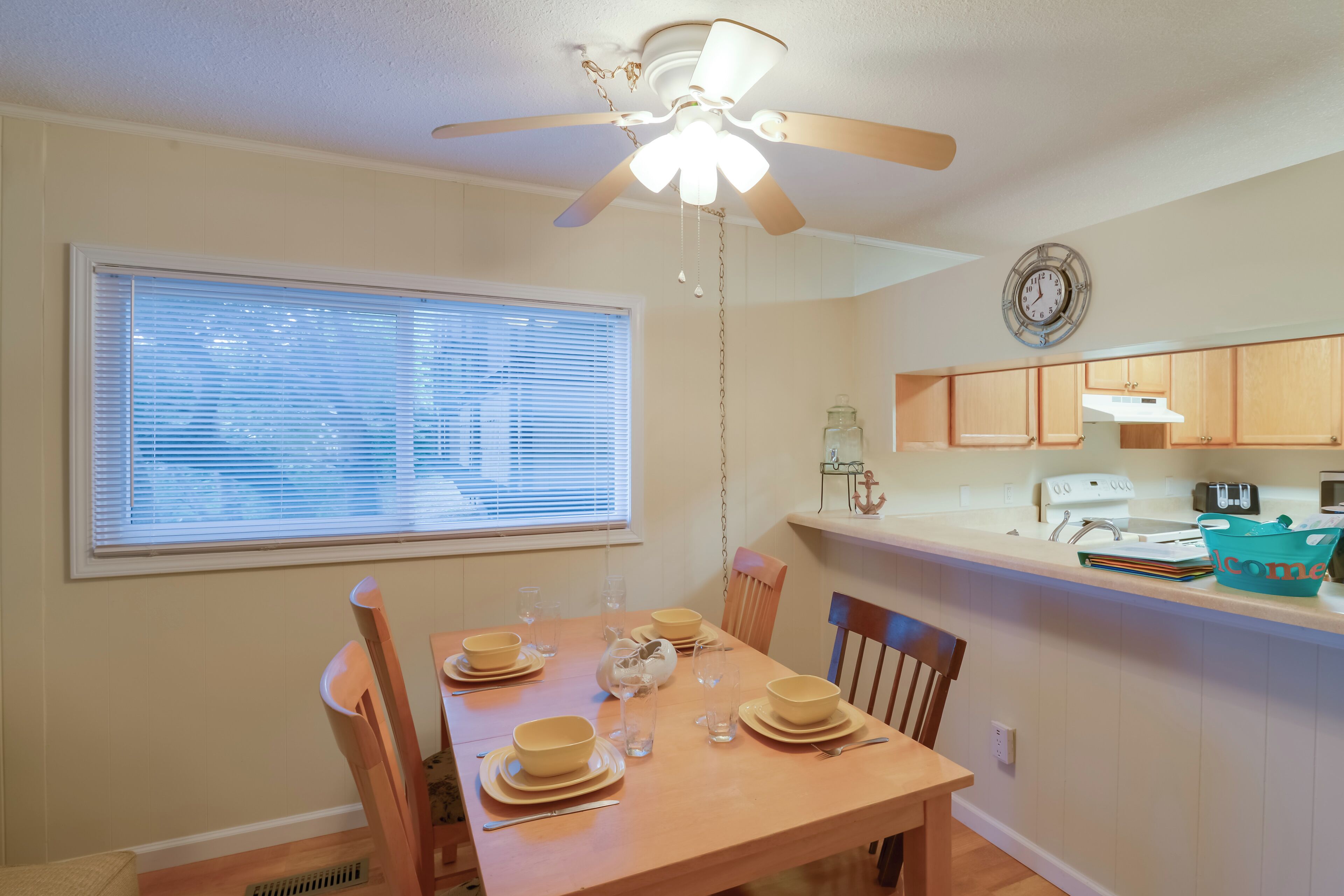 A dining area with a wooden table set for four, a ceiling fan, and a window with blinds.