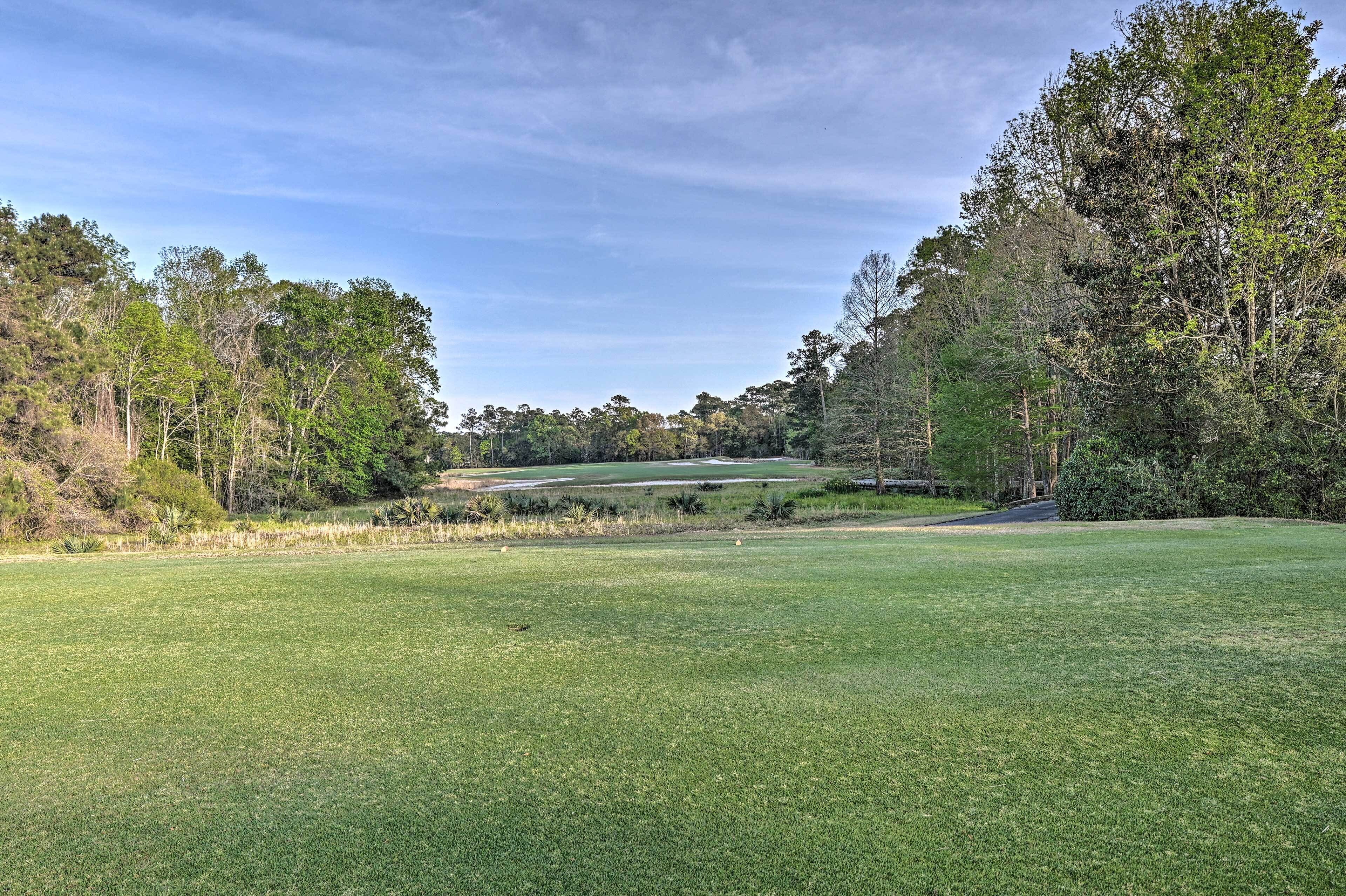 A golf course with a well-maintained green, surrounded by dense trees and a clear sky.