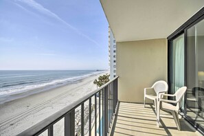 A balcony with a view of the beach, two white plastic chairs, and a glass door. - Oceanfront Vacation Rental in Murrells Inlet! (Murrells Inlet)