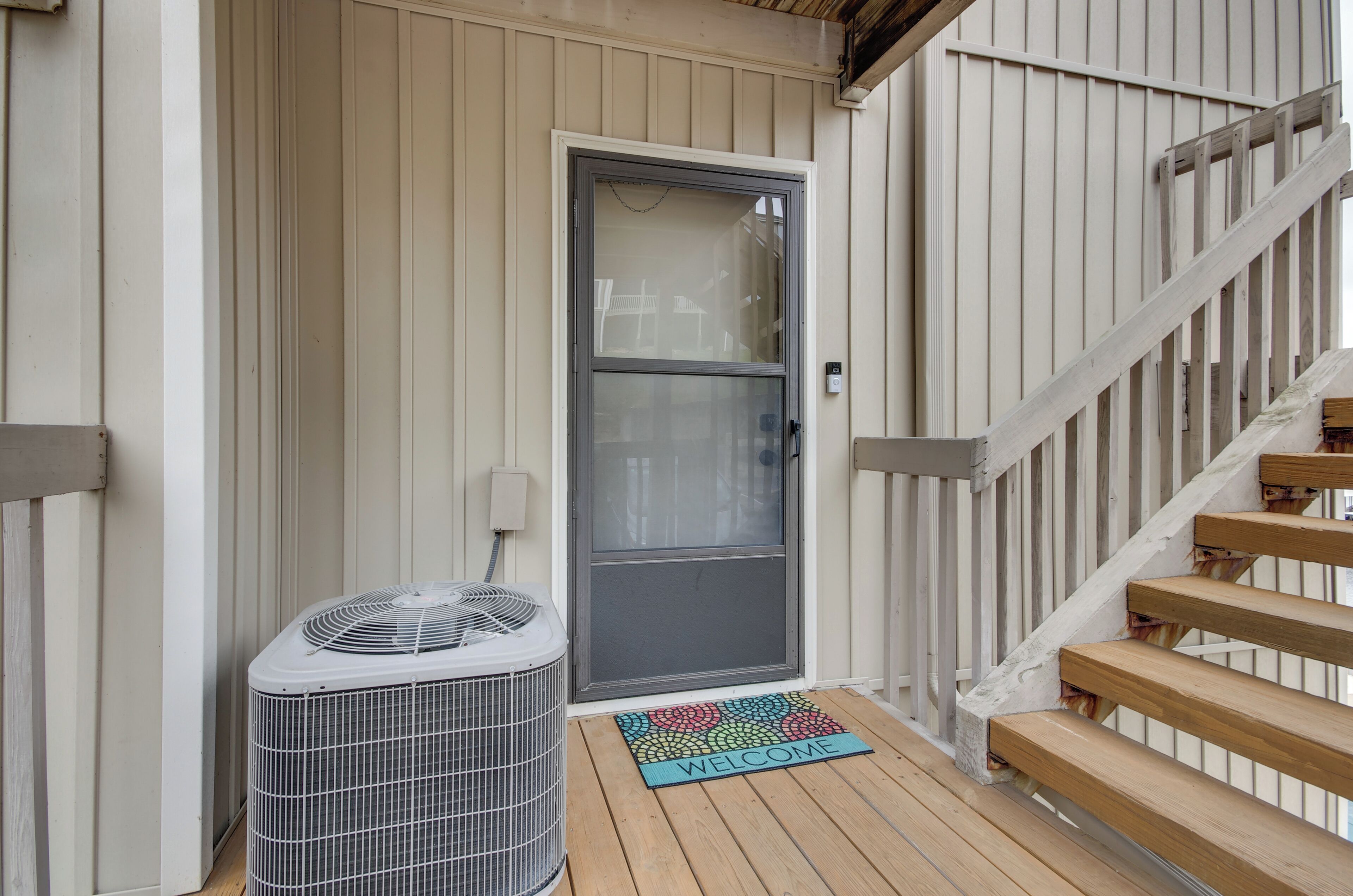 En træterrasse med velkomstmåtte, en grå dør med glasrude, og en stående aircondition.