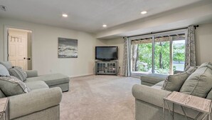 A spacious living room with two grey sofas, a flat-screen TV on a cabinet, a large window with curtains, and a door leading to another room. - Dock, Slide + Fire Pit: Osage Beach Lake House (Osage Beach)