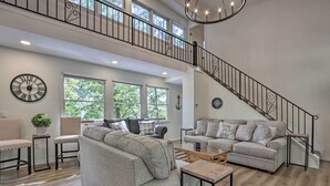A spacious living room with a high ceiling, a staircase, and large windows. - Dock, Slide + Fire Pit: Osage Beach Lake House (Osage Beach)