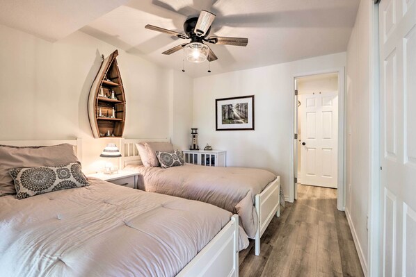 A bedroom with two beds, a ceiling fan, a wooden boat-shaped shelf, a framed picture, and a door leading to another room.