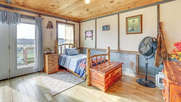 Une pièce aux murs lambrissés, avec un lit, une commode en bois, un ventilateur et une vue par la fenêtre.