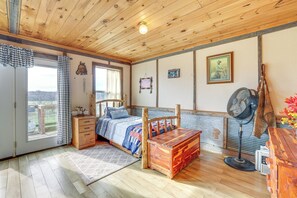 Une pièce aux murs lambrissés, avec un lit, une commode en bois, un ventilateur et une vue par la fenêtre.