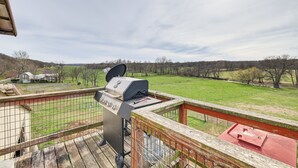 Une terrasse dotée d’un barbecue et d’une structure surmontée d’un toit rouge, offrant une vue sur un champ verdoyant et des bâtiments situés au loin.