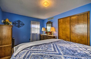 A bedroom with a blue wall, a wooden wardrobe, a bed with a patterned bedspread, a nightstand with lamps, and a mirror. - Peaceful Missouri Home, 25 Mi to Truman Lake! (Cole Camp)