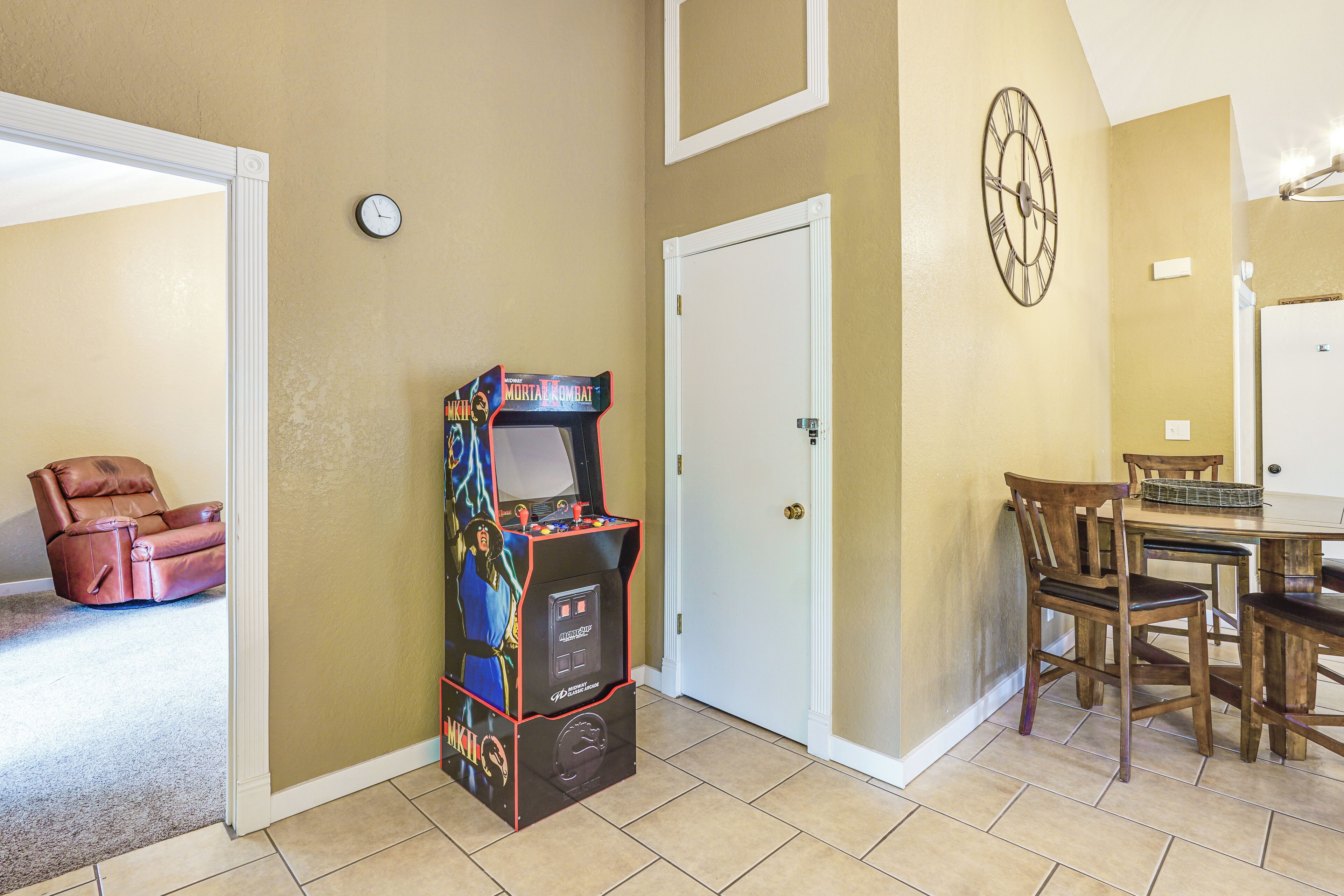 Une pièce comprenant une console de jeux, une table à manger avec des chaises, une horloge et une porte.