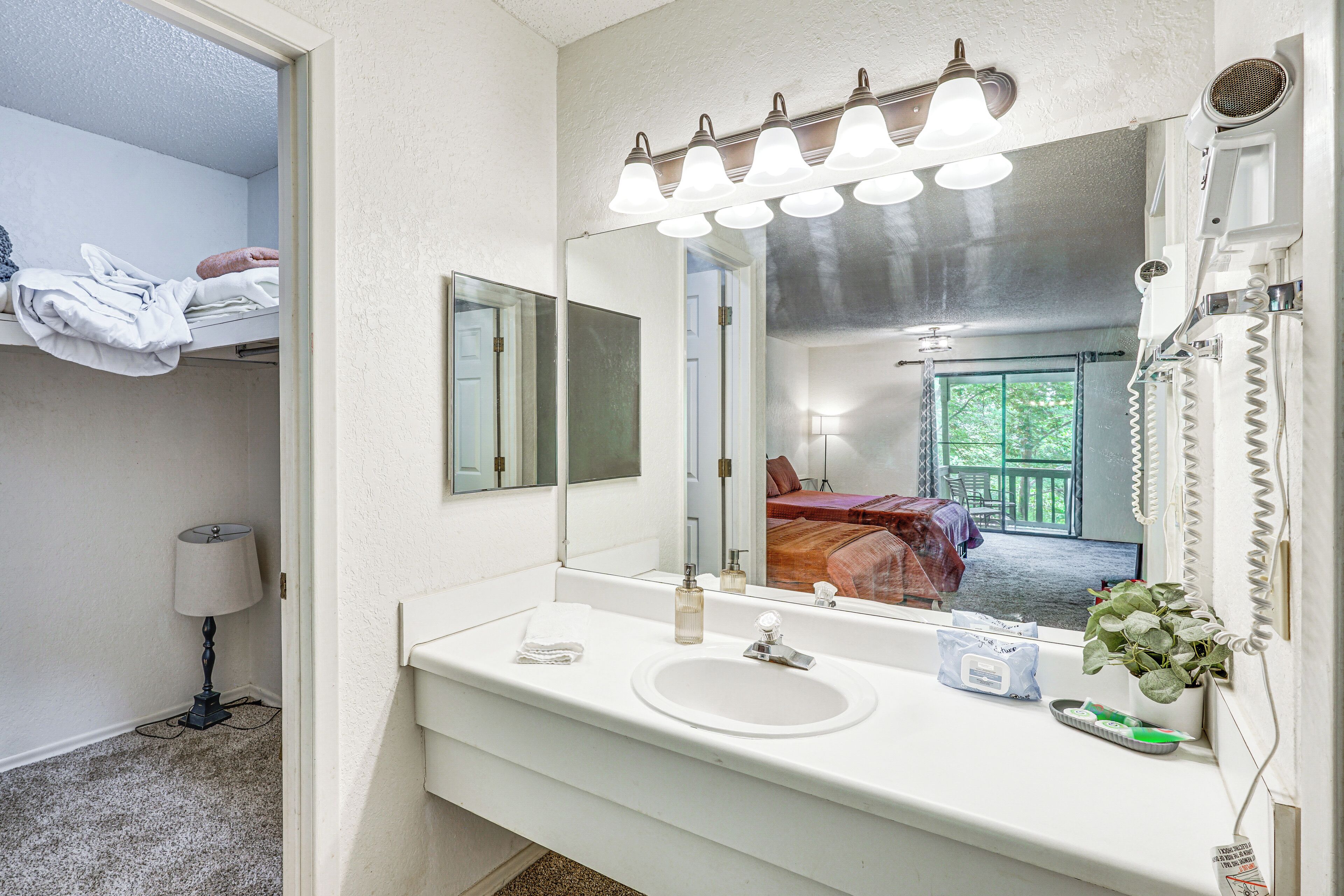 A bathroom with a white sink, mirror, and vanity. There is a towel rack above the sink, a lamp, and a window with a view of a room with a bed.