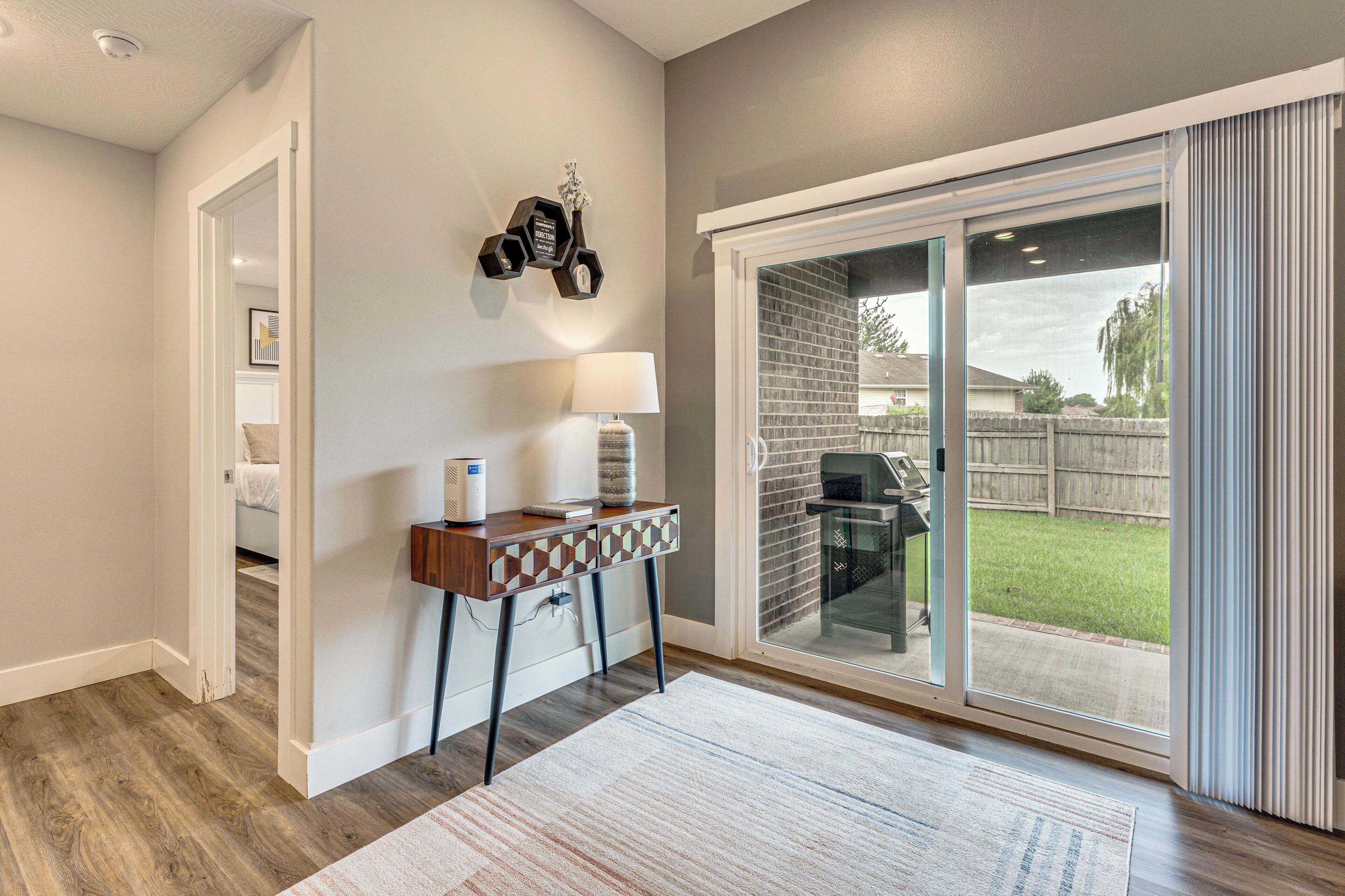 A modern interior with a wooden floor, a desk with a lamp, and a sliding glass door leading to a backyard.