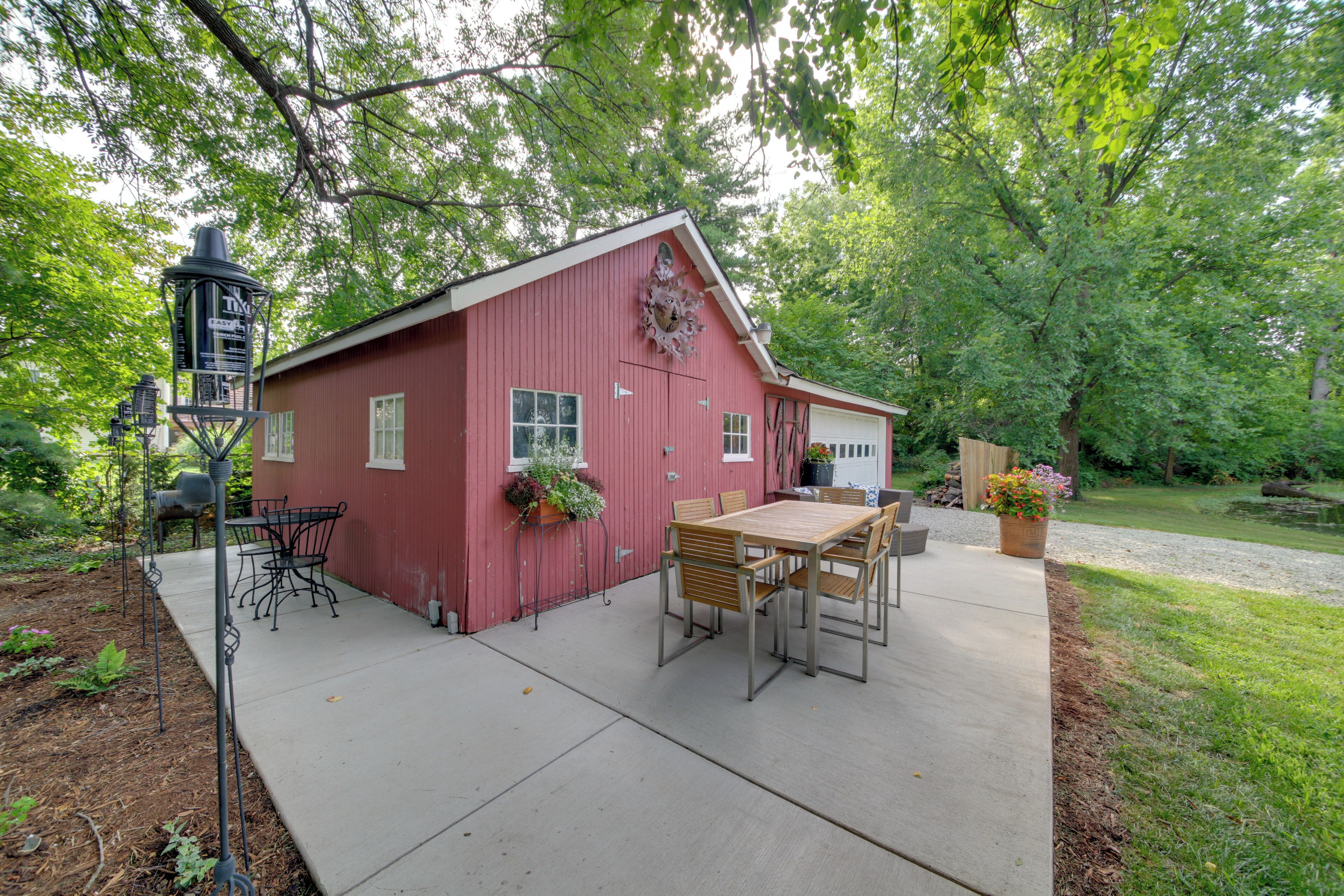 A red barn with a wooden table and chairs outside, surrounded by greenery.
