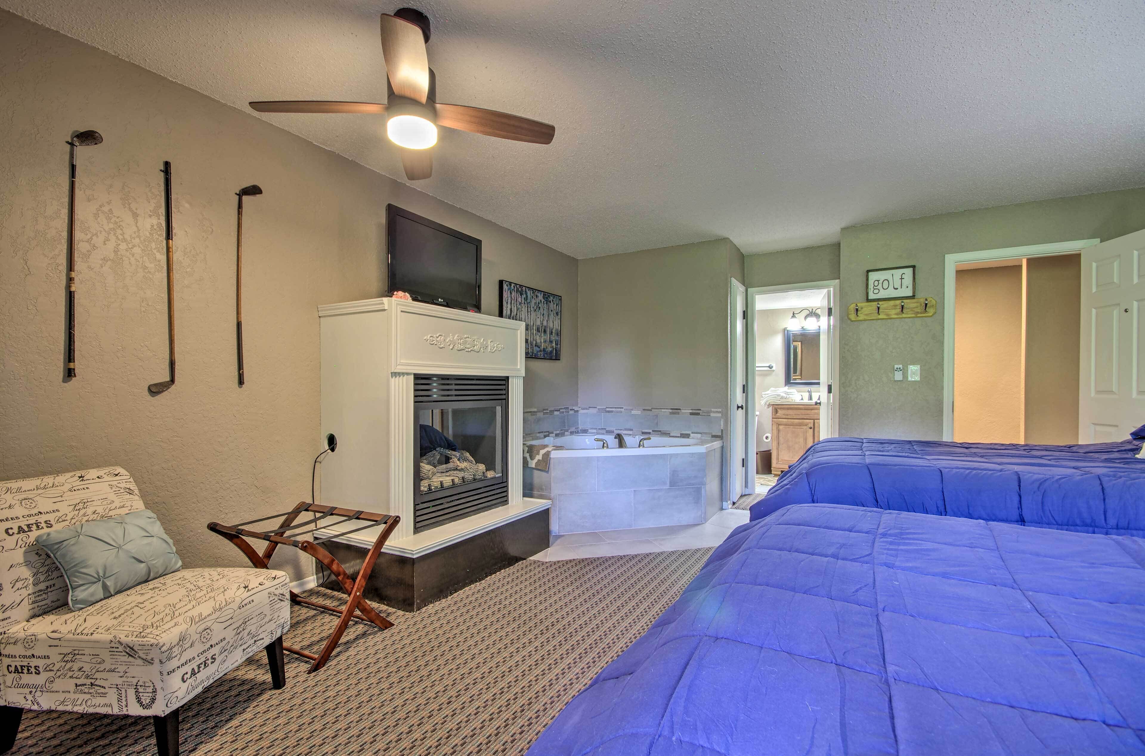 A bedroom with a large bed, a fireplace, a ceiling fan, and a bathroom visible in the background.
