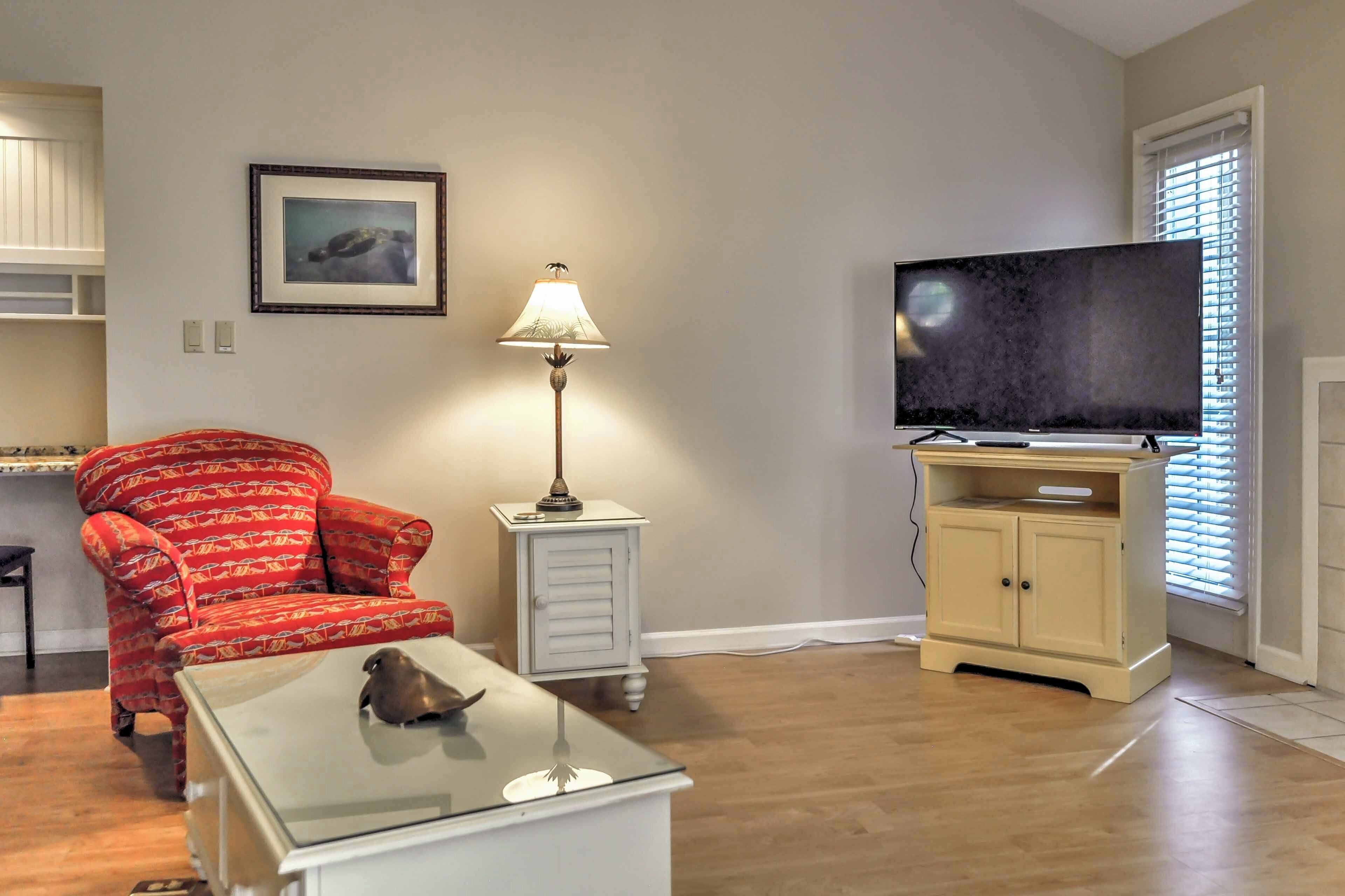 A living room with a red patterned armchair, a glass coffee table with a bird sculpture, a white side table with a lamp, a television on a stand, and a beige cabinet.