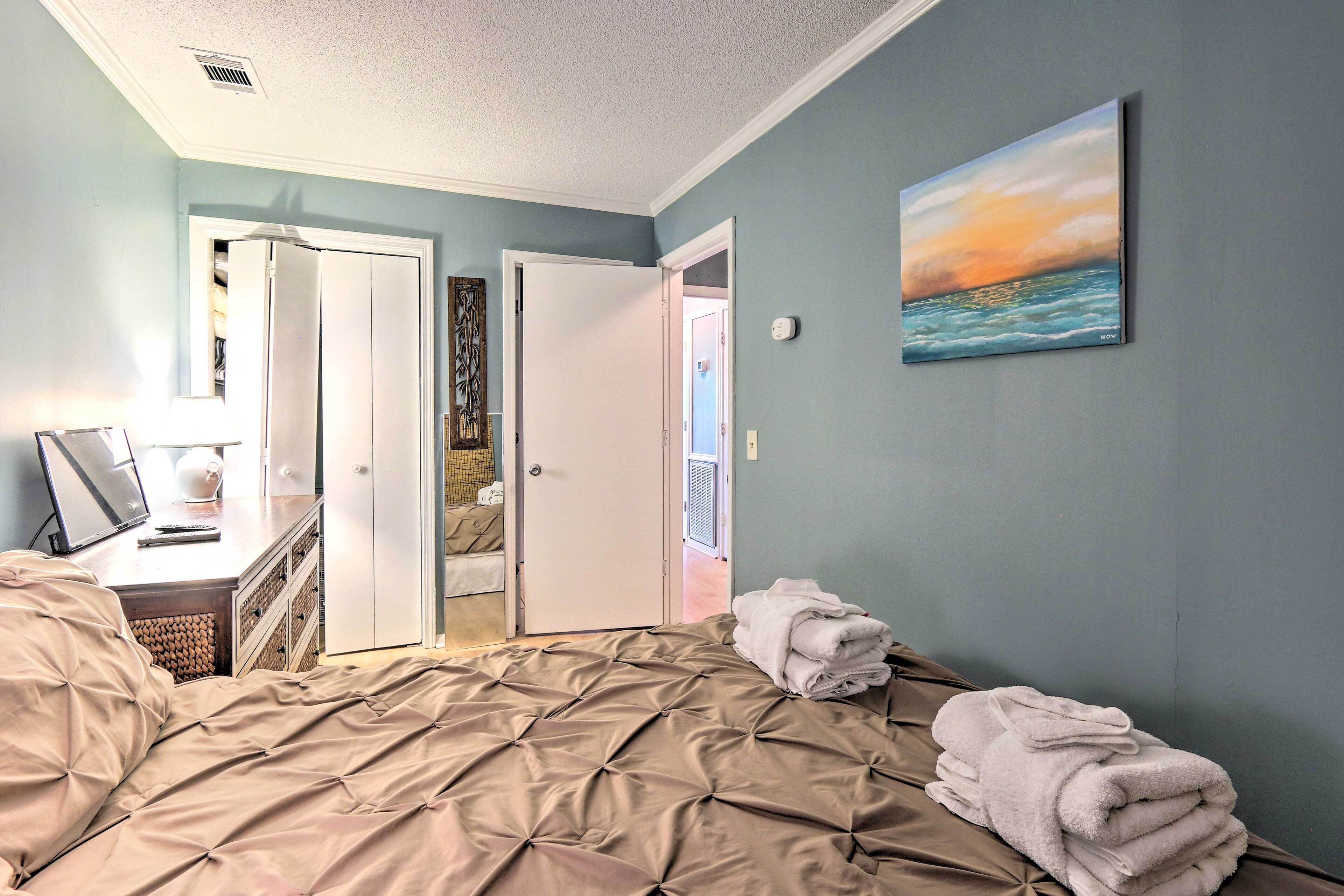 A bedroom with a bed, a desk, a computer, a painting, and a door leading to another room.