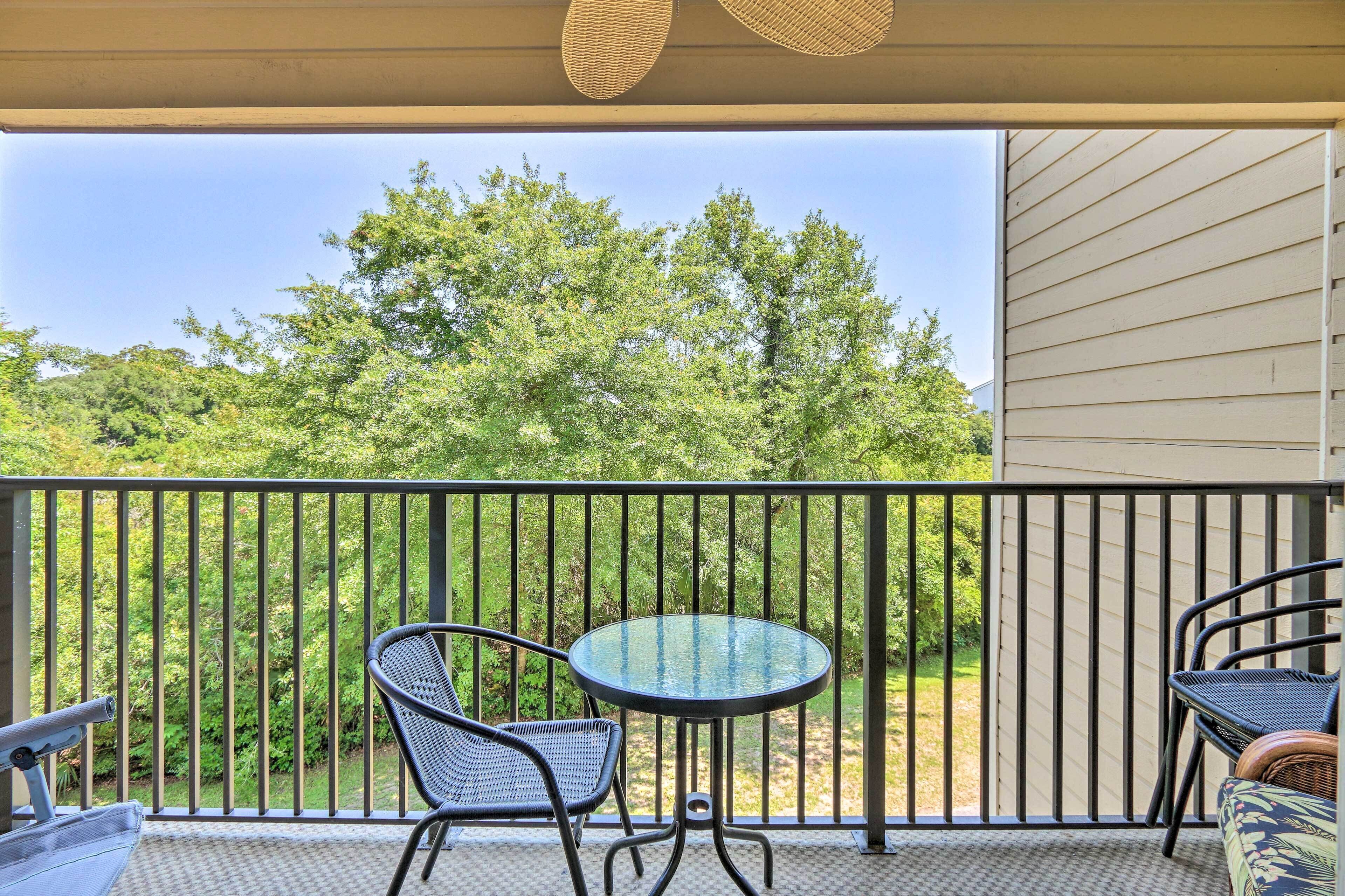A balcony with a glass table and two chairs, overlooking a garden with trees.