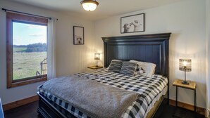 A bedroom with a large bed, two bedside lamps, a window with a view of a field, and a framed picture on the wall. - Rustic Missouri Vacation Rental w/ Hill Views! (Greenfield)