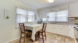 Una cocina con armarios blancos, una mesa de comedor con sillas y un ventanal con cortinas blancas.