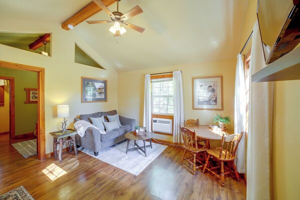A cozy living room with a sofa, coffee table, and dining table. There is a ceiling fan, a window with curtains, and a door leading to another room.