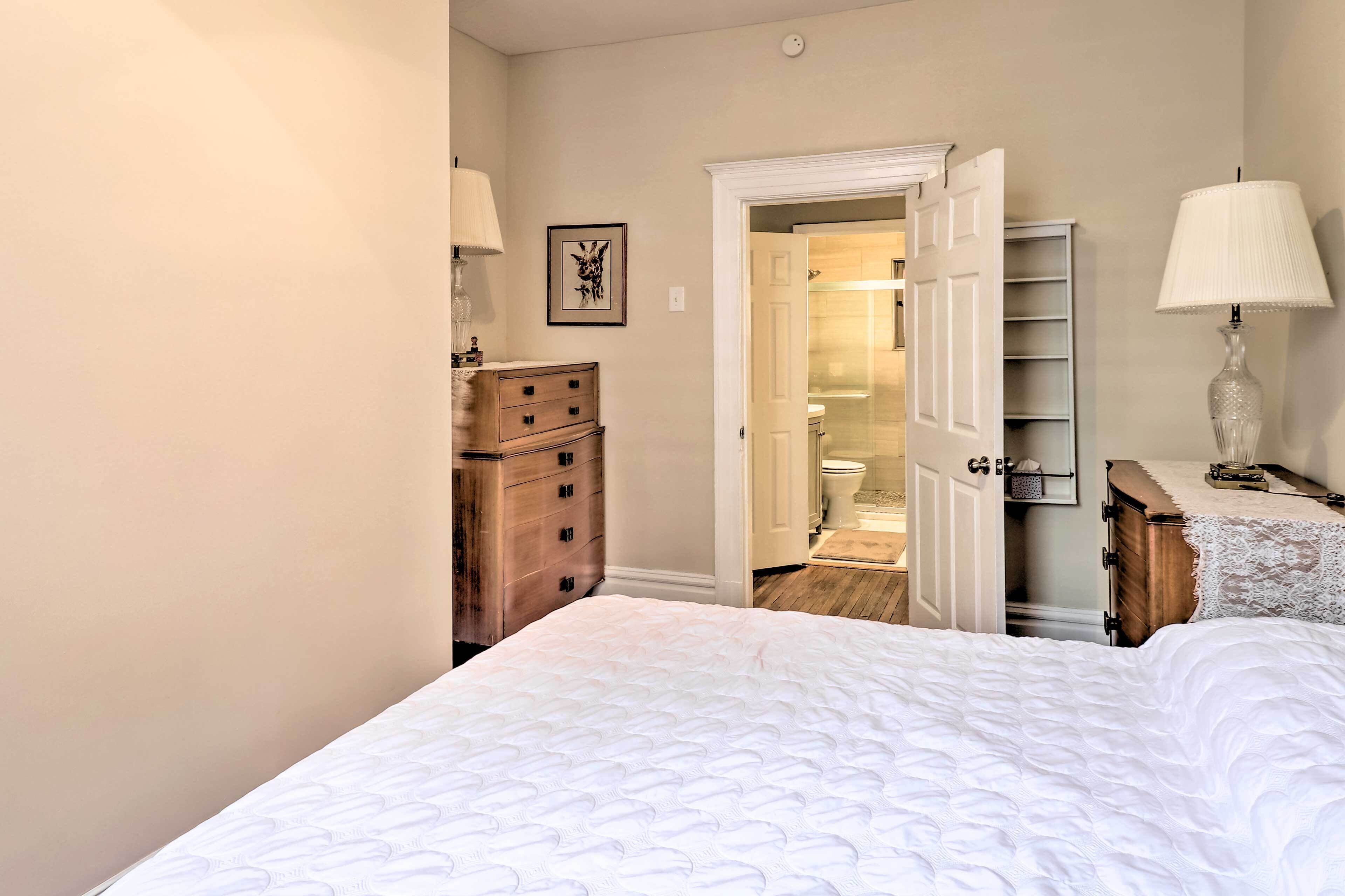 A bedroom with a large bed, a wooden dresser, a bedside table with a lamp, and a door leading to a bathroom.