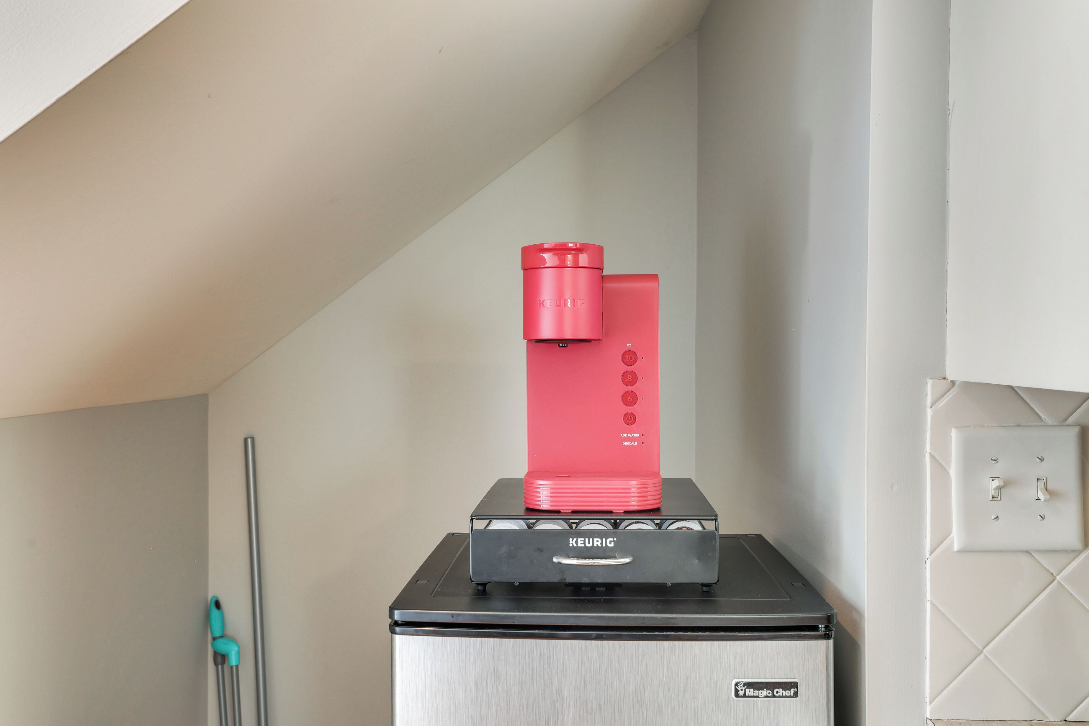 A kitchen with a coffee maker on a black countertop, a teal-handled mop, and a white tiled wall.