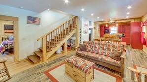 A spacious living area with a wooden staircase, a sofa with patterned cushions, and a kitchen area with red cabinets. - Rustic Hollister Cabin - 5 Mi to Downtown Branson (Hollister)