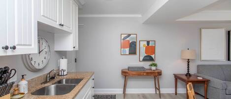 A modern kitchen with white cabinets, a granite countertop, stainless steel sink, and a clock. There is a small wooden table with a lamp and a gray couch in the living area.