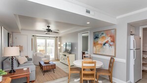 A modern living room with a sofa, coffee table, and dining table with chairs. There is a ceiling fan, a TV, and a refrigerator in the background. - Summerhouse Villas Condo: Private Resort Beach! (Pawleys Island)