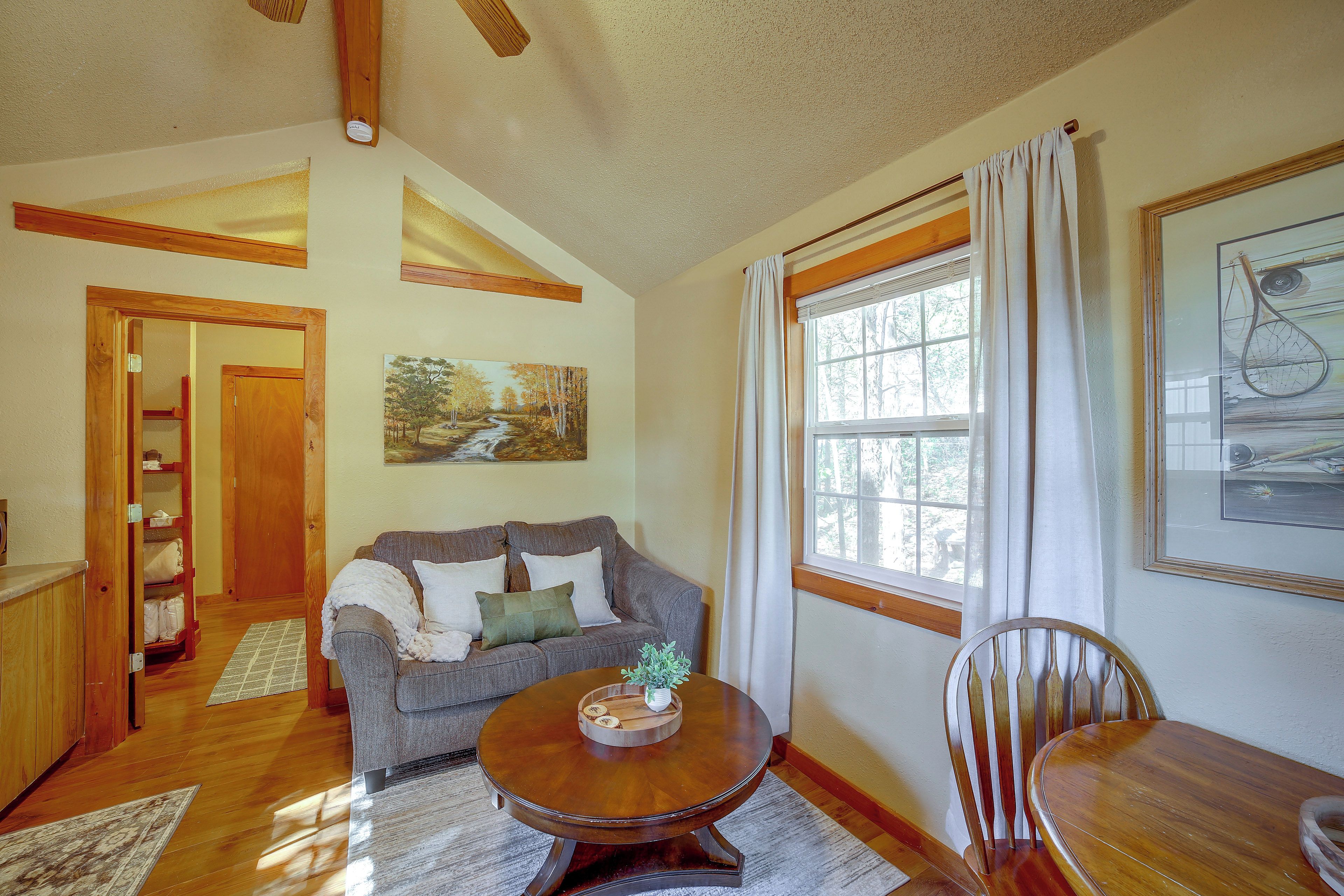 A cozy living room with a wooden floor, a grey sofa, a round wooden table, and a window with curtains.