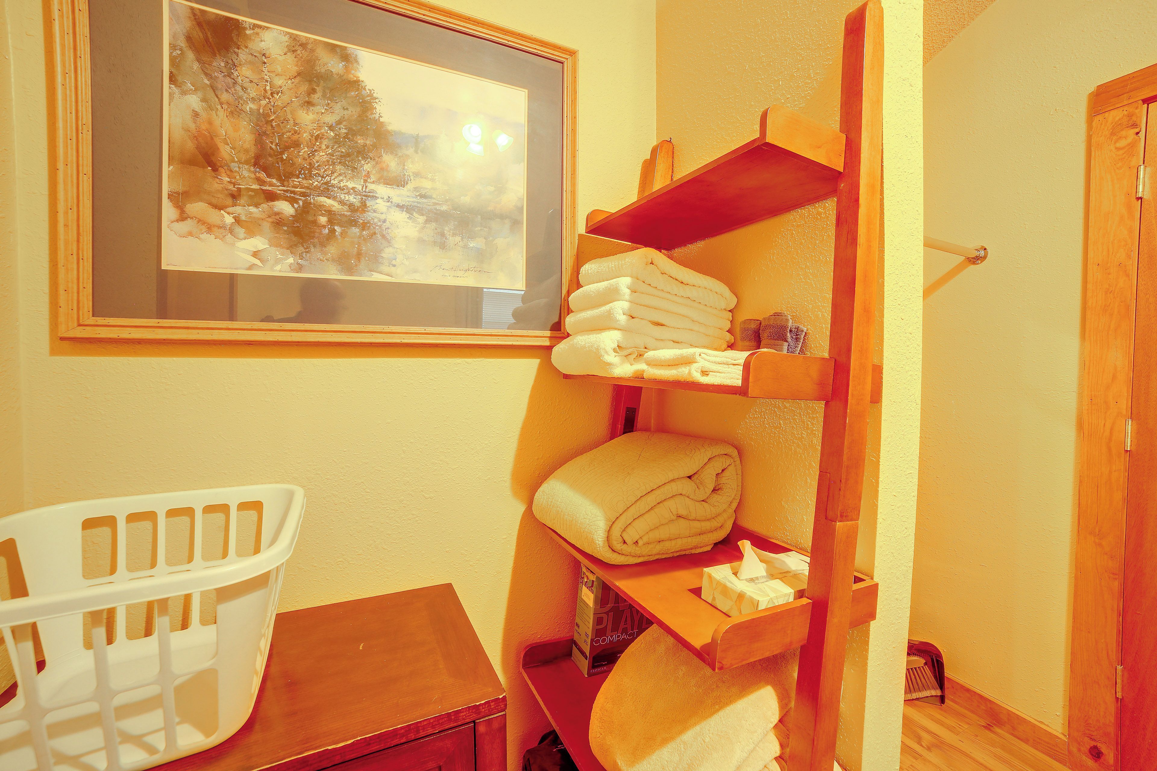 A bathroom with a wooden shelf holding towels, a framed painting on the wall, and a white plastic chair.