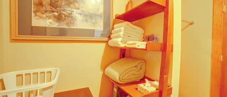 Un baño con un estante de madera que sostiene toallas, un cuadro en la pared y una silla de plástico blanco.