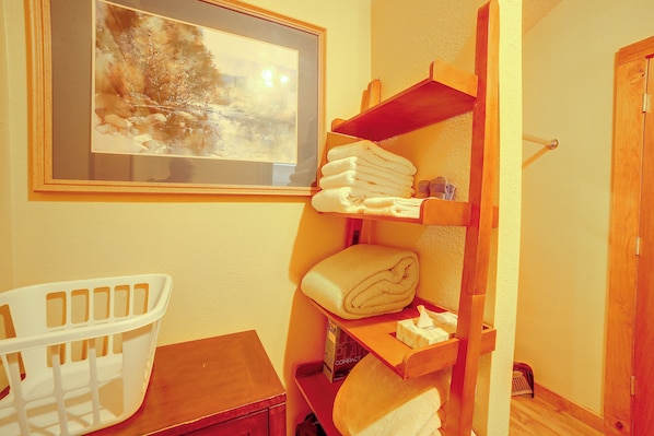 A bathroom with a wooden shelf holding towels, a framed painting on the wall, and a white plastic chair.