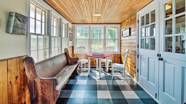 A cozy room with a wooden ceiling, large windows, a checkered floor, a bench, and a table with chairs. - Remodeled Historic Farmhouse ~ 20 Mi to KC! (Liberty)