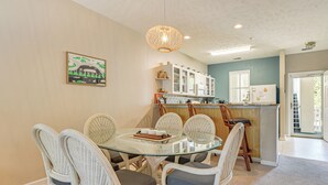 A dining area with a glass table surrounded by wicker chairs, a kitchen with white cabinets, and a hanging light fixture. - Sunny Coastal Condo ~ 1 Mi to N Myrtle Beach! (North Myrtle Beach)
