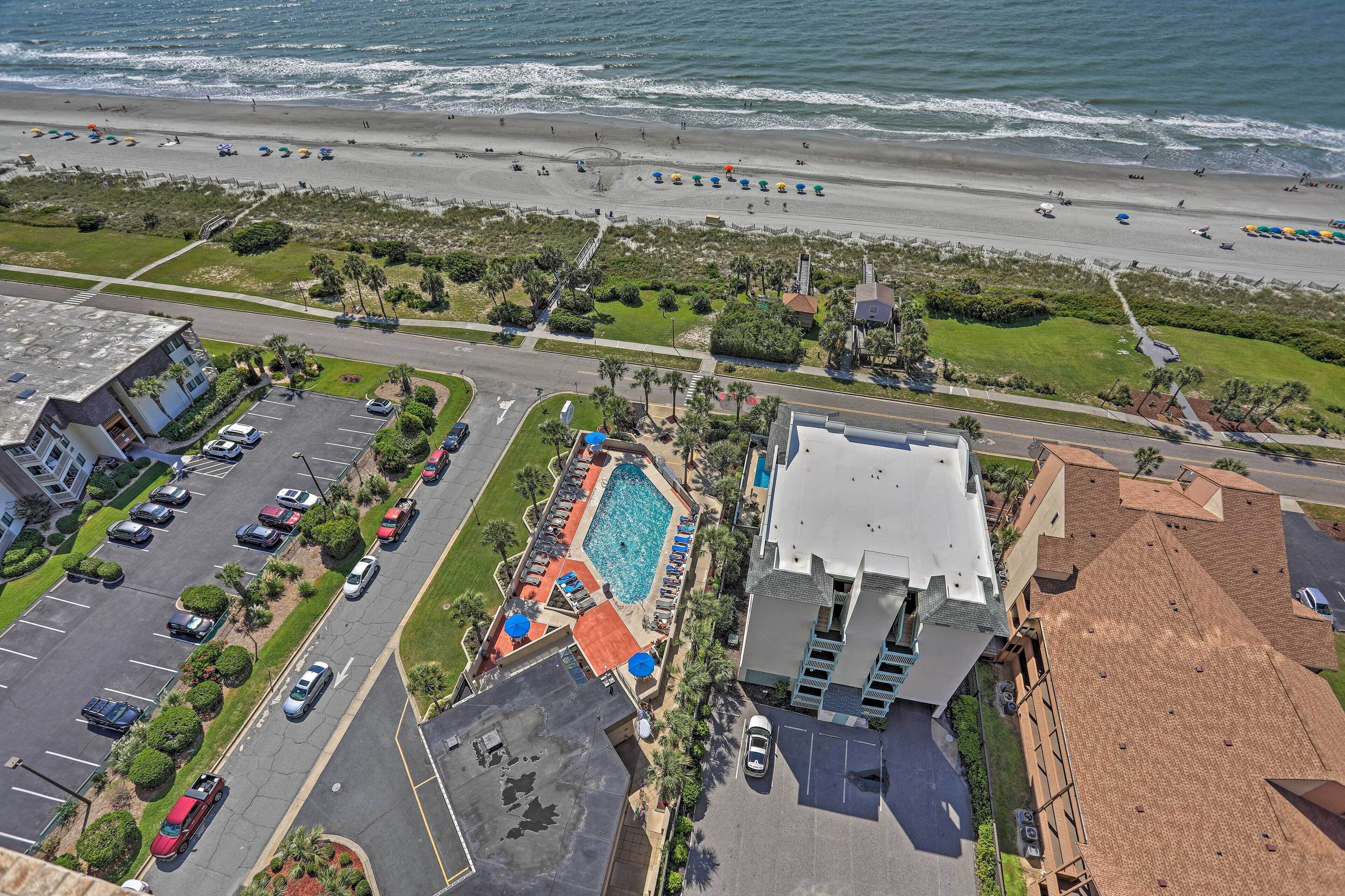 An aerial view of a coastal hotel with a swimming pool, parking lot, and beach access.