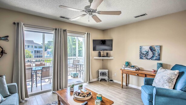 A living room with a ceiling fan, a TV mounted on the wall, a blue armchair, a wooden coffee table with books and a remote, and a sliding glass door leading to an outdoor patio. - Sunny N Myrtle Condo w/ Pool < 2 Mi to Beach! (North Myrtle Beach)