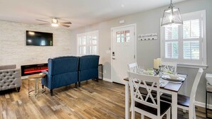 Un salon moderne doté d’une cheminée, d’un téléviseur fixé au mur en briques, d’un ventilateur de plafond et d’un coin repas avec une table dressée pour quatre personnes.