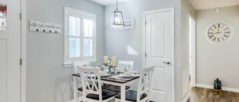 A dining area with a wooden table and white chairs, a hanging light fixture, and a wall clock.