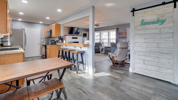 A modern kitchen with wooden cabinets, a dining area with a wooden table and benches, and a living room with a large window and a television. - Table Rock Lake Home - Fire Pit & Lake Views! (Golden)